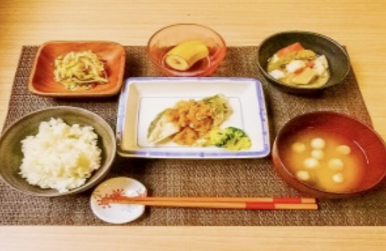 米飯、味噌汁(ミズナ・麩)、鮭の白身魚の葱ソース、里芋の味噌煮、竹輪の胡麻和え、果物(バナナ)